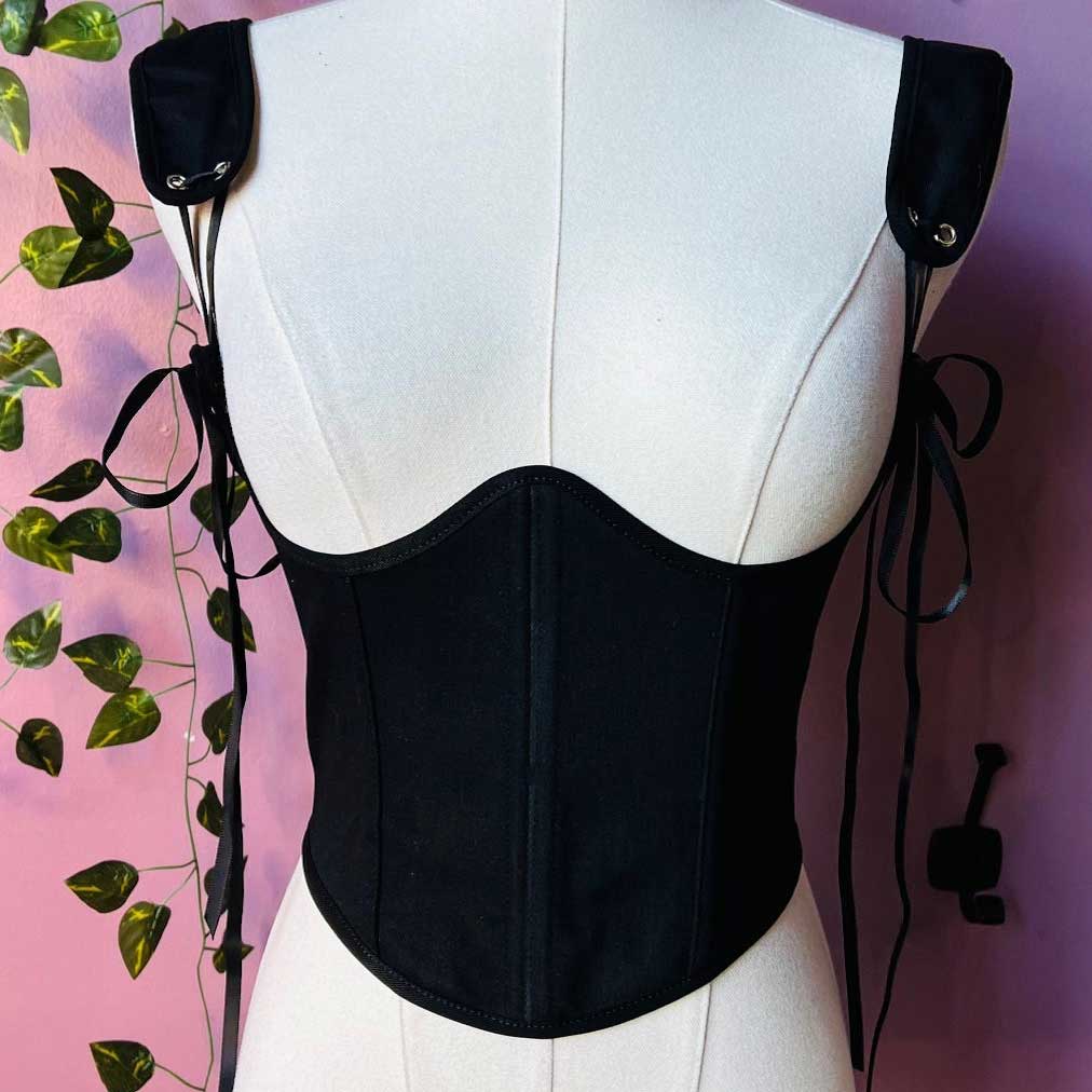 Black corset on a mannequin against a pink background with green leaves.